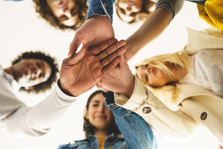 A photo of a group of people stood in a circle from below, with their hands together in the middle of the circle.