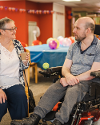 An elderly lady sat next to a man in an electric wheelchair in a community hall decorated with balloons and party decorations.